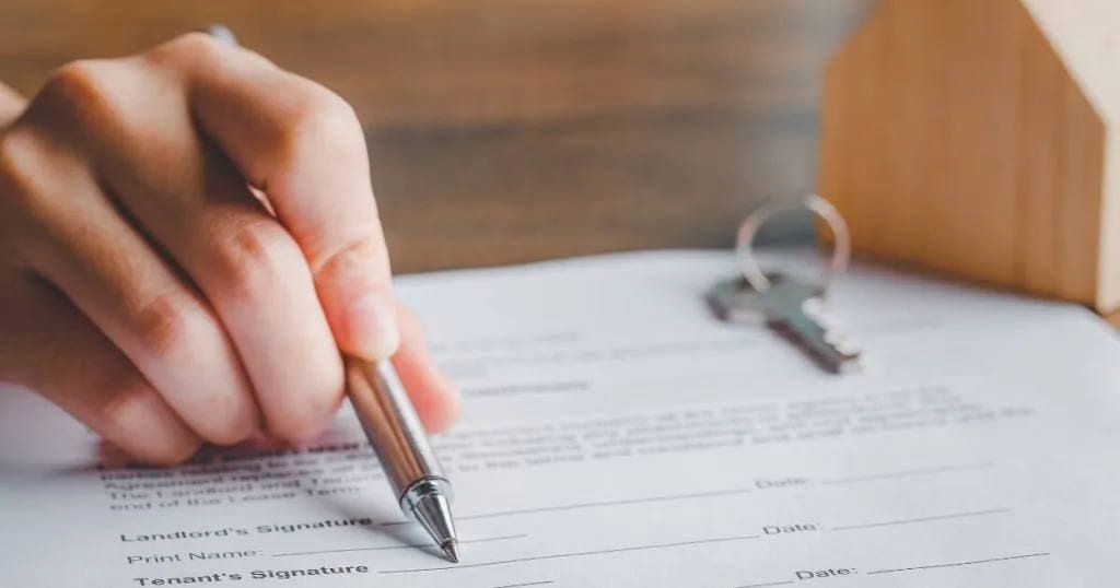 Hand signing a lease agreement beside a set of keys and a small wooden house