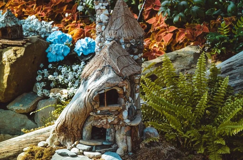Small stone and driftwood fairy house in a garden bed with ferns and hydrangeas
