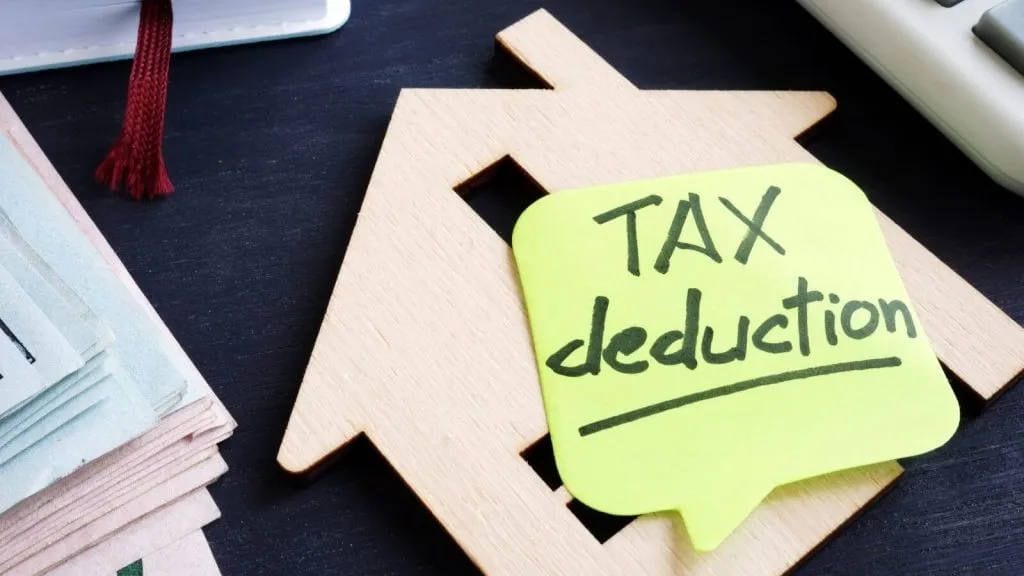 Sticky note reading Tax deduction on a wooden house cutout beside stacked cash